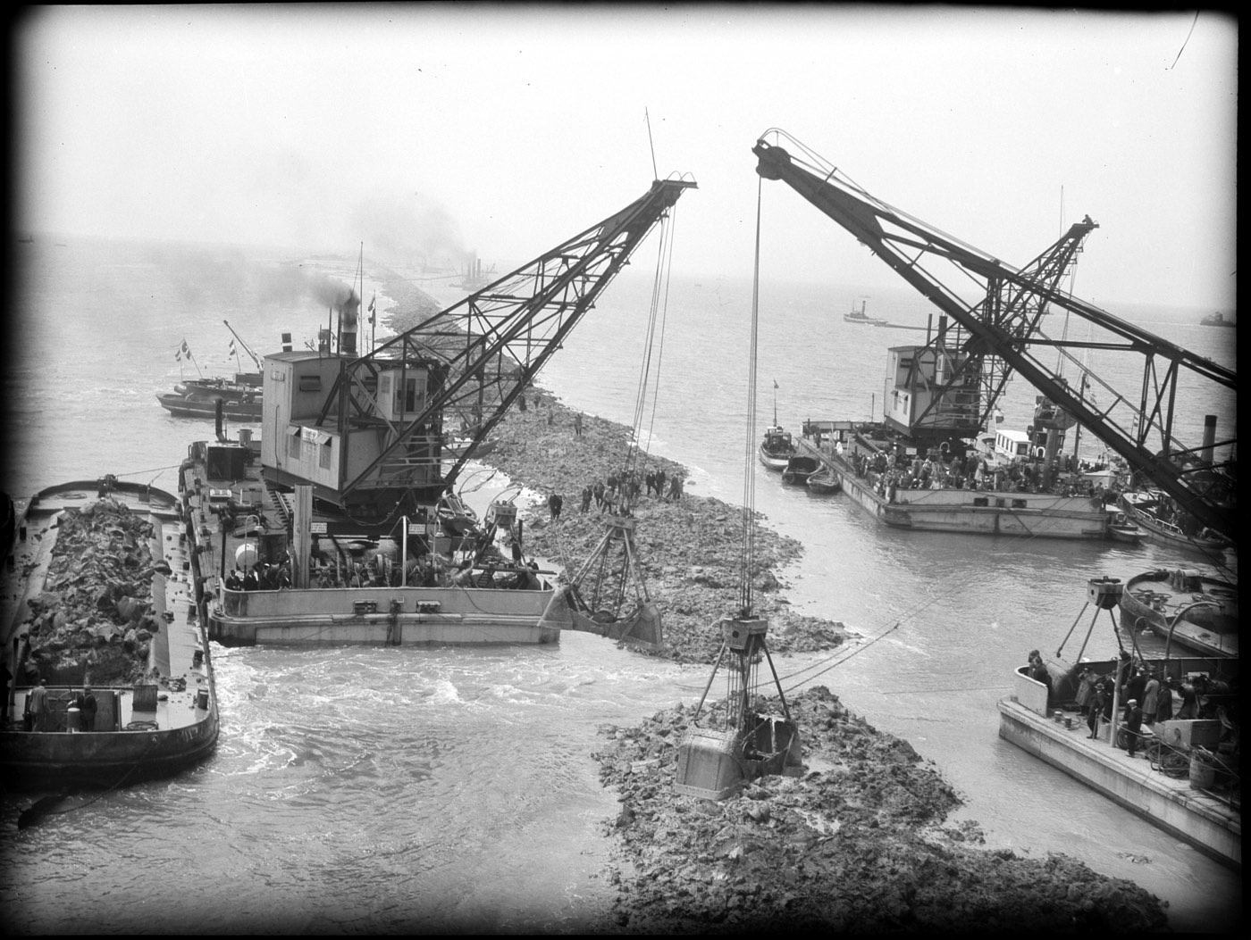 Black-and-white photograph from 1932 of floating cranes and clay barges closing De Vlieter, the final gap in the Afsluitdijk, with crowds watching from the decks
