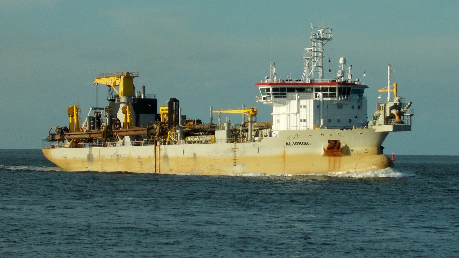 The trailing suction hopper dredger Al-Idrisi under way in open water, suction pipes stowed along her yellow-craned hull