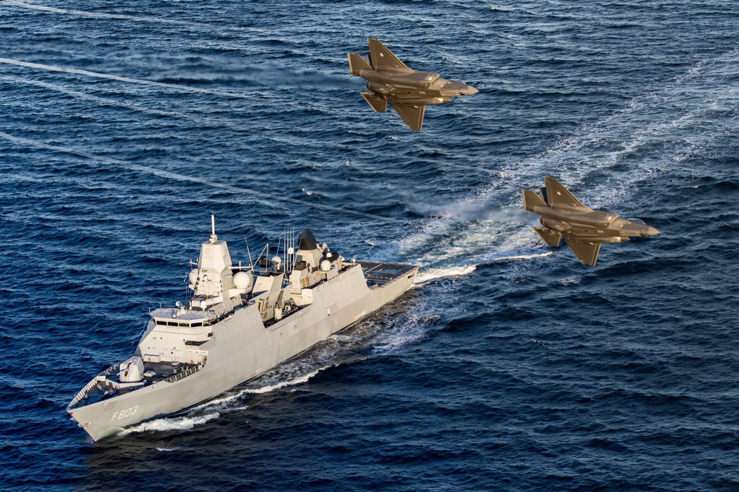 The Dutch frigate Zr.Ms. Tromp under way in open water as two F-35 fighters pass low overhead
