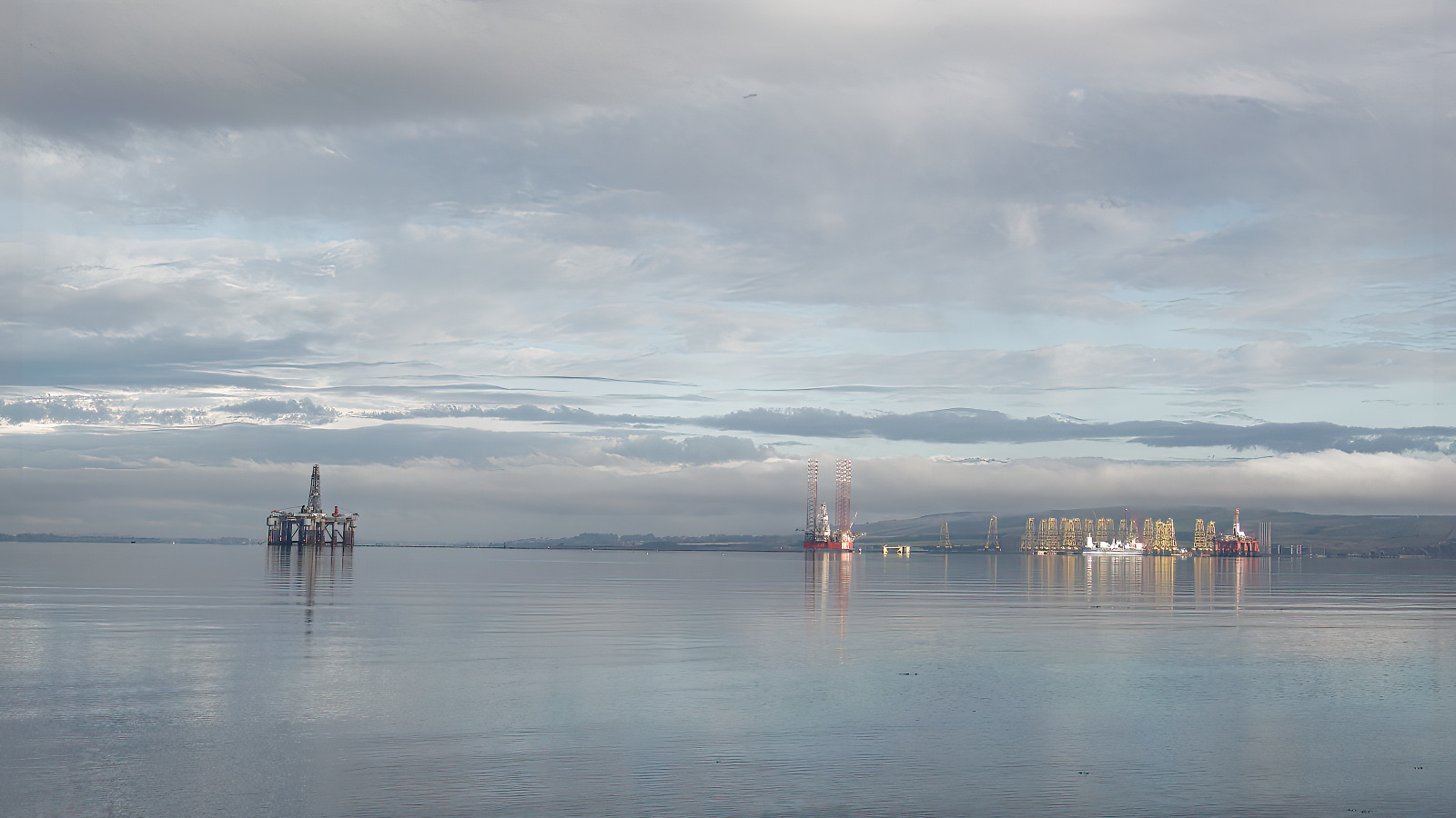 A wide, calm Cromarty Firth under grey cloud, with a semi-submersible rig at anchor in the foreground and a cluster of stacked jack-up rigs lit by low sun along the far shore