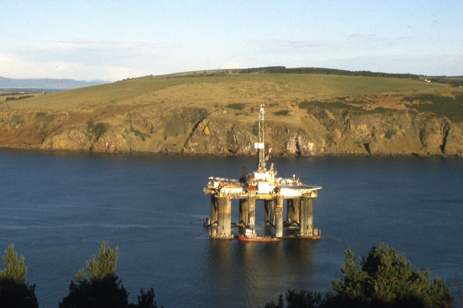 A semi-submersible drilling rig under tow through the narrow entrance of the Cromarty Firth, tugs alongside, with the cliffs of the Sutors behind