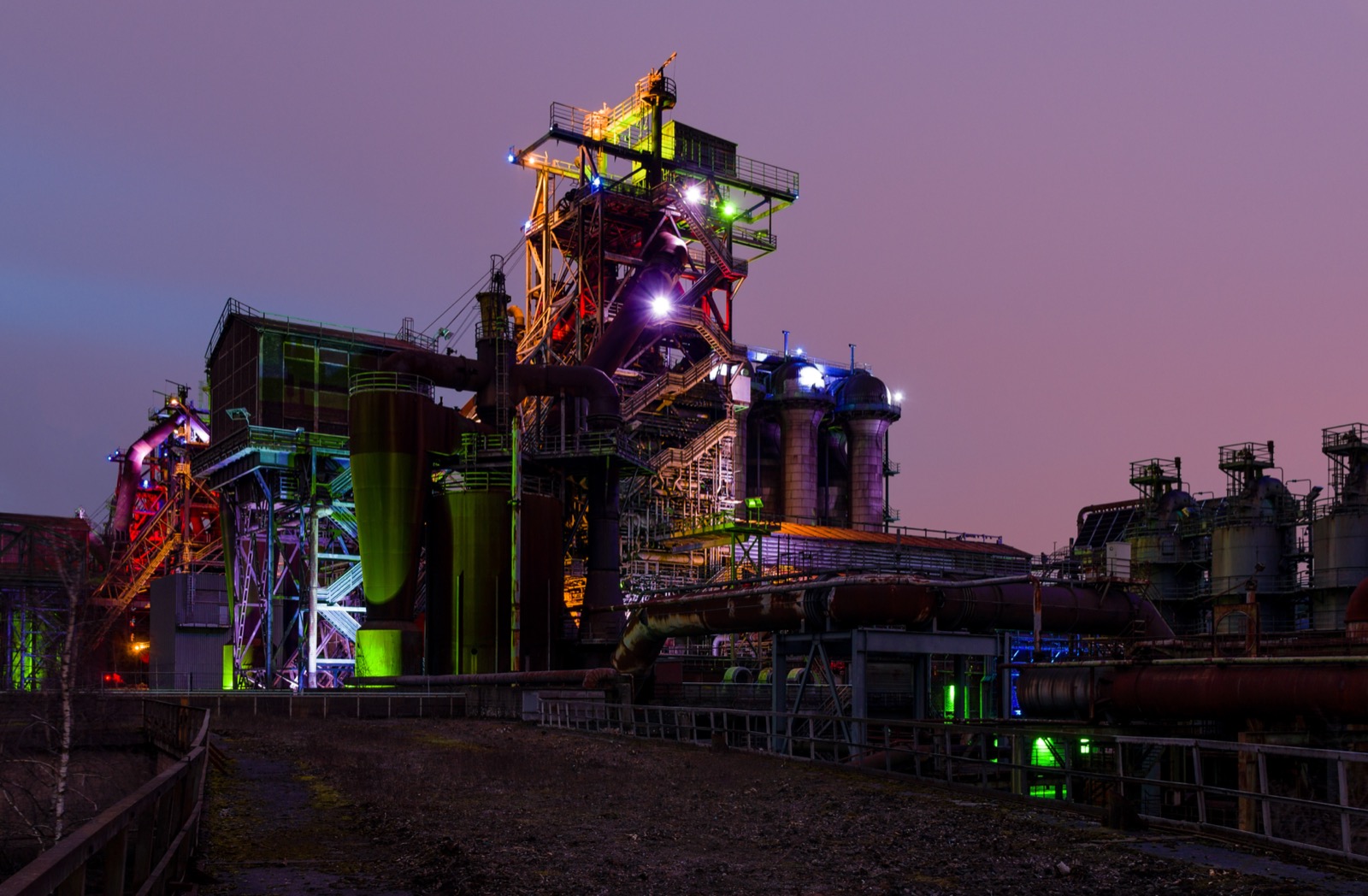 A disused blast furnace complex at Landschaftspark Duisburg-Nord lit in vivid colours at dusk, with walkways and pipework picked out against a purple sky