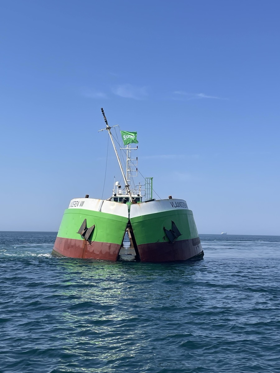 A green and red barge seen head-on at sea with its hull split open down the centre line, the two halves hinged apart above the waterline