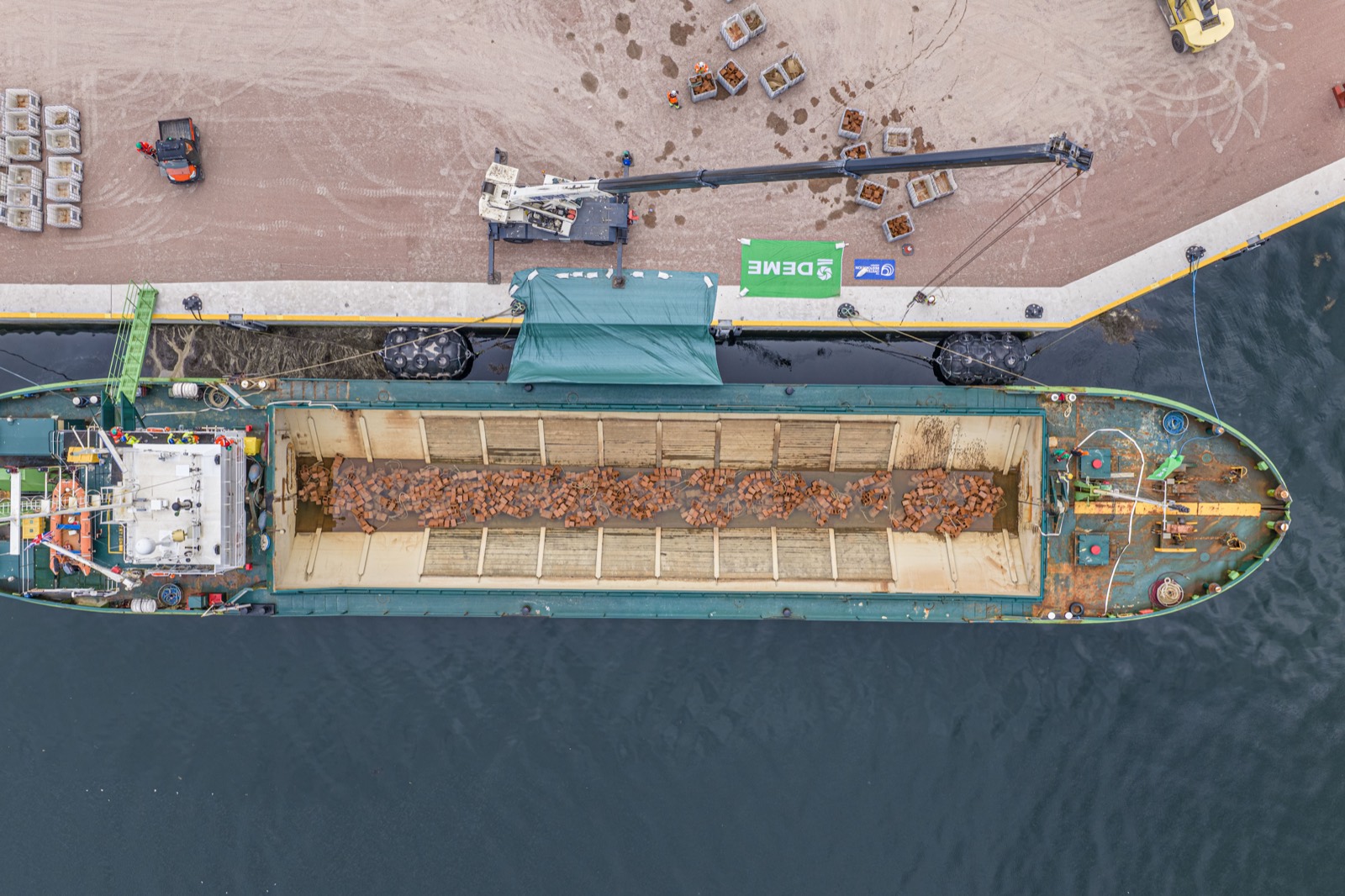 Overhead drone view of a green split hopper barge alongside a quay, its long hopper loaded with terracotta clay bricks, with a crane and stillages of bricks on the dockside