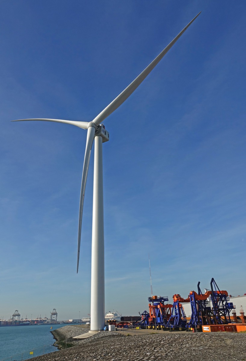 A single very large offshore wind turbine standing on a rock revetment against a clear blue sky, dwarfing the port cranes and equipment beside it