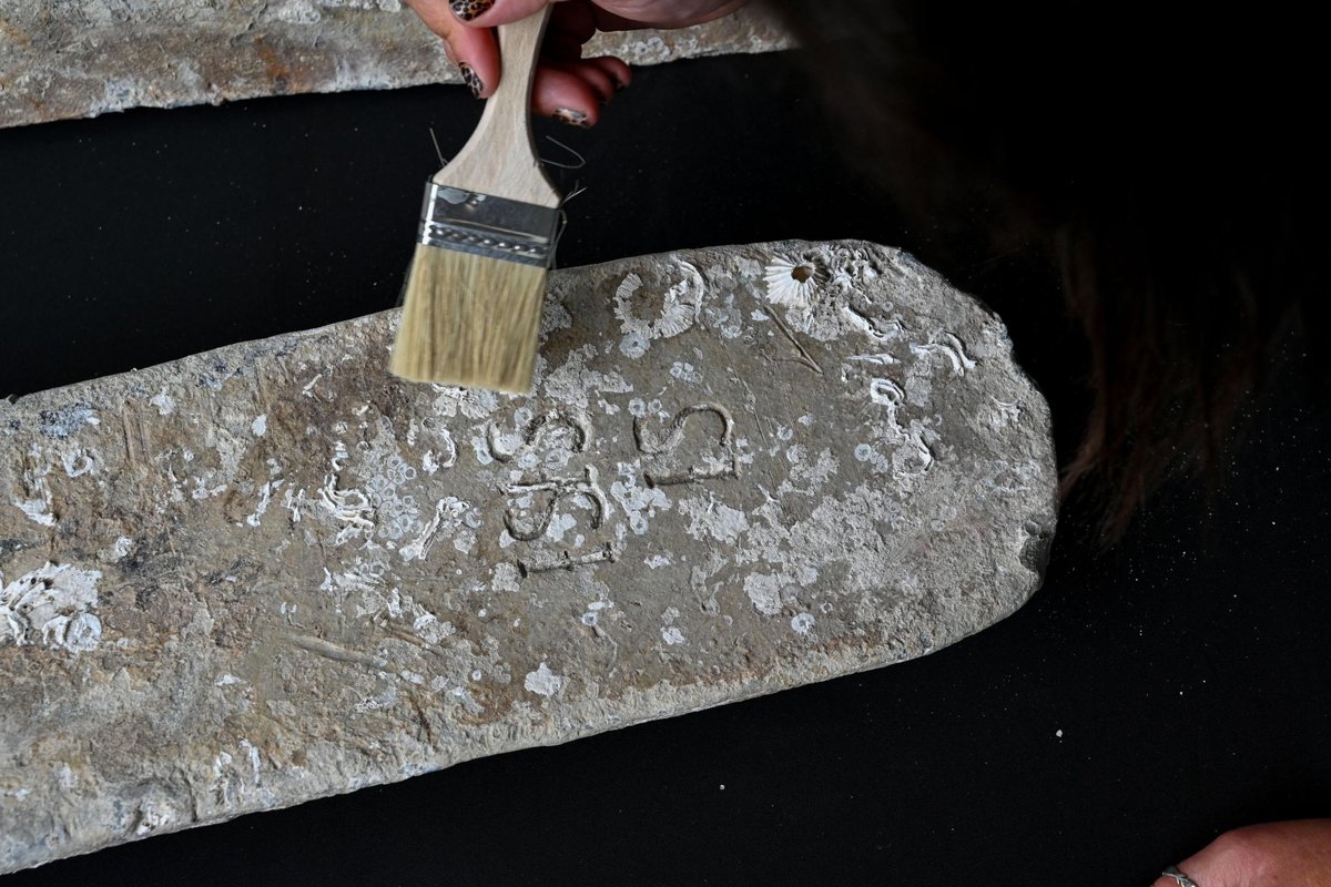 A hand working a brush across an encrusted lead ingot, the stamped maker's mark showing through the marine growth