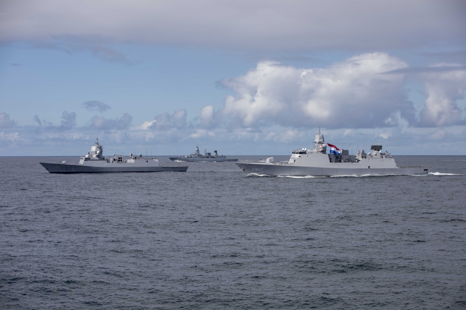 Three NATO frigates, Norwegian, German and Dutch, under way on open grey water beneath a high cloudbank