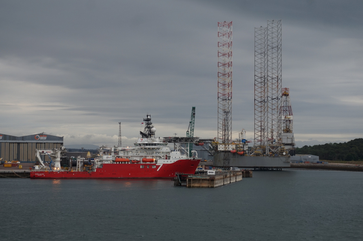 A jack-up drilling rig with its legs raised alongside a red offshore support vessel at the quay at Nigg, under heavy grey cloud