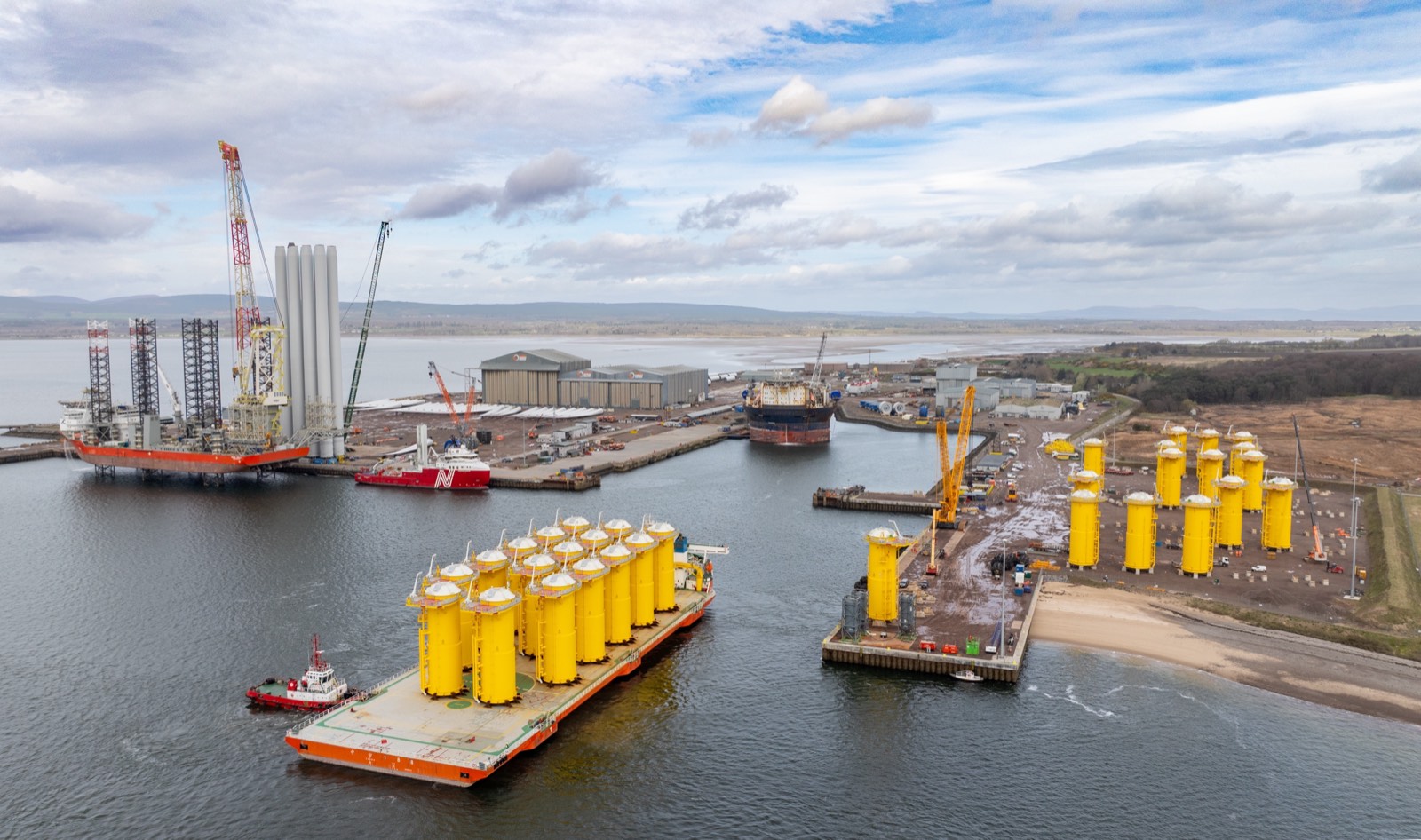 Aerial view of Maraen Port of Nigg: a barge loaded with yellow offshore wind transition pieces under tow, more stacked on the quay, a jack-up vessel and fabrication sheds behind