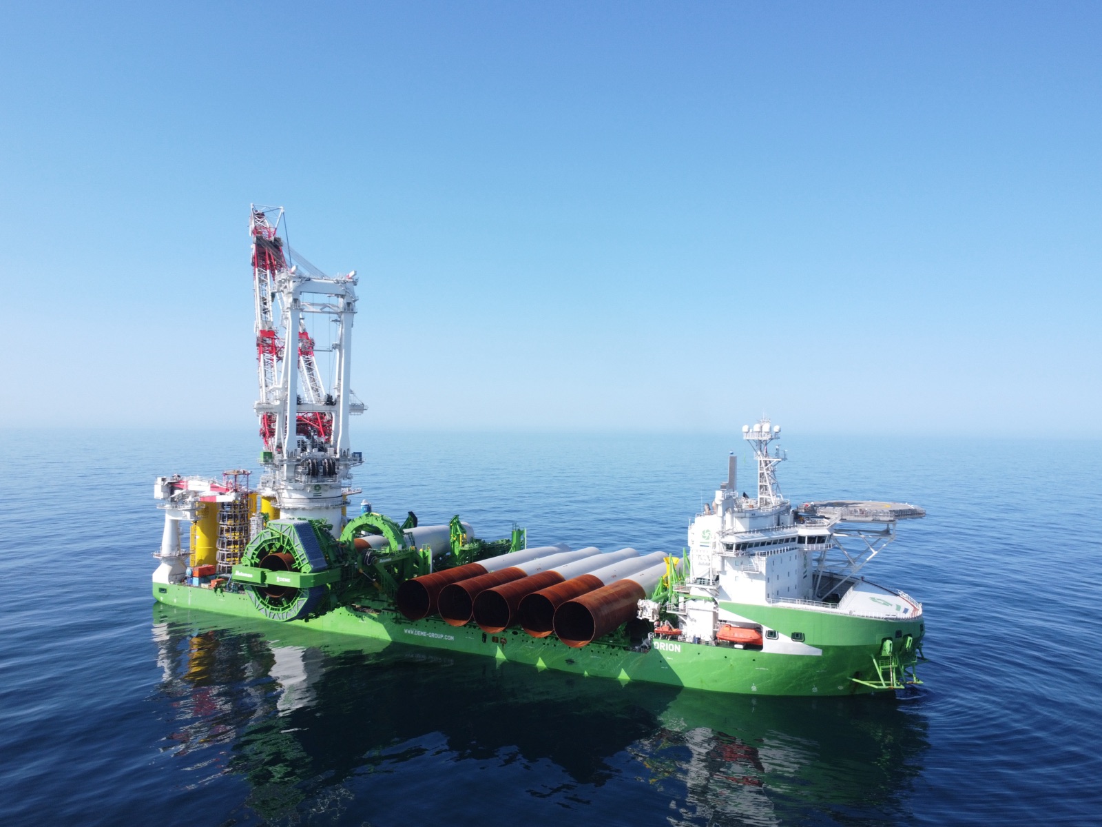 Aerial view of DEME's offshore installation vessel Orion in a calm sea, five rust-brown monopile foundations stacked on deck beneath the towering crane
