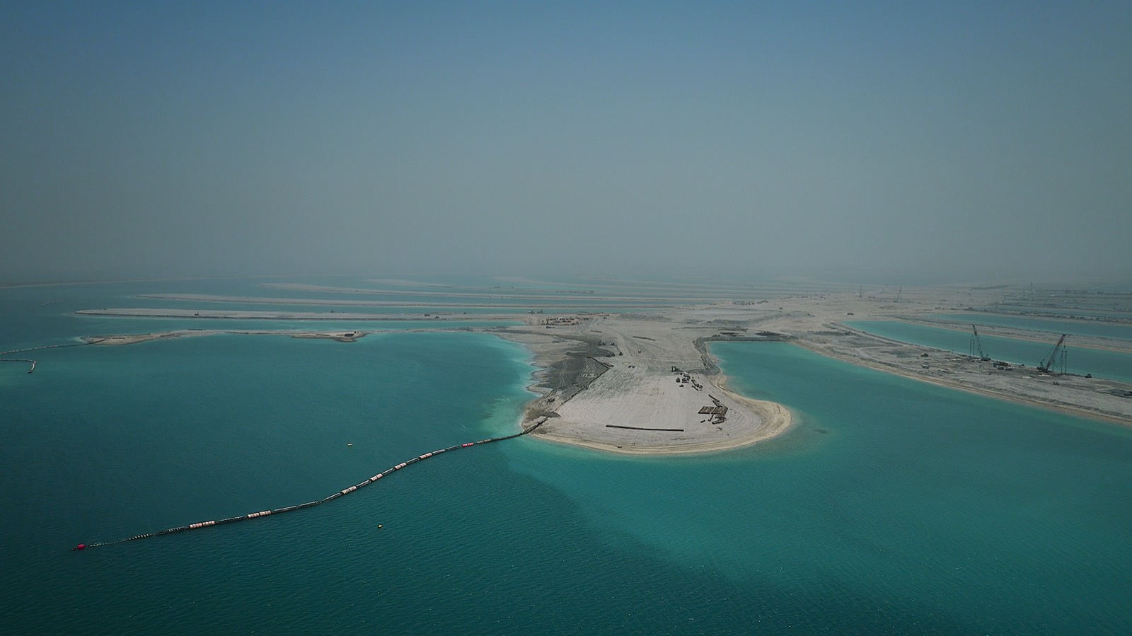 Aerial view of a frond of Palm Jebel Ali under construction: pale new land in turquoise water, with a black floating pipeline running out to sea