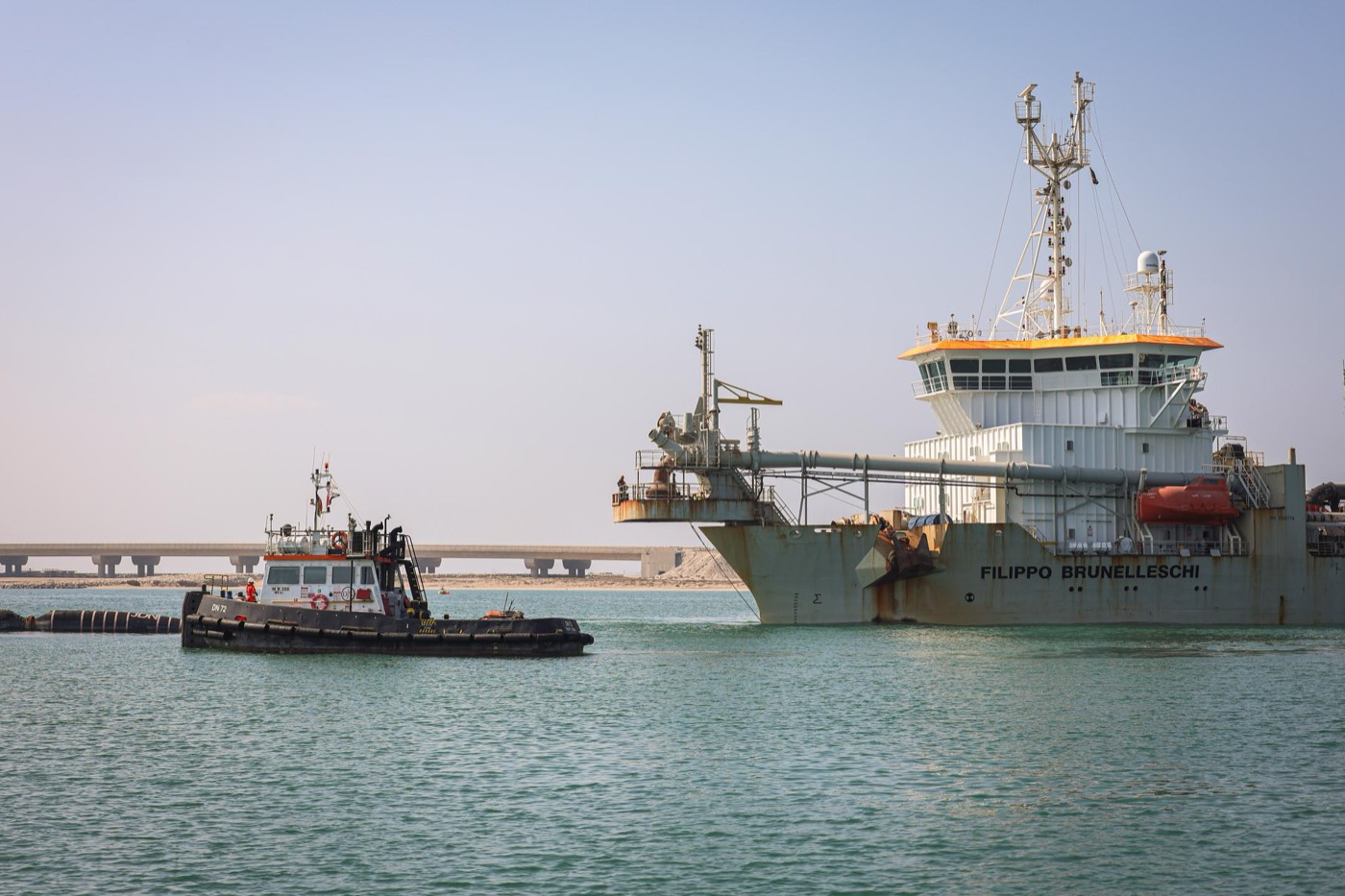 The trailing suction hopper dredger Filippo Brunelleschi lying off the reclamation site with a tug alongside and a bridge causeway behind