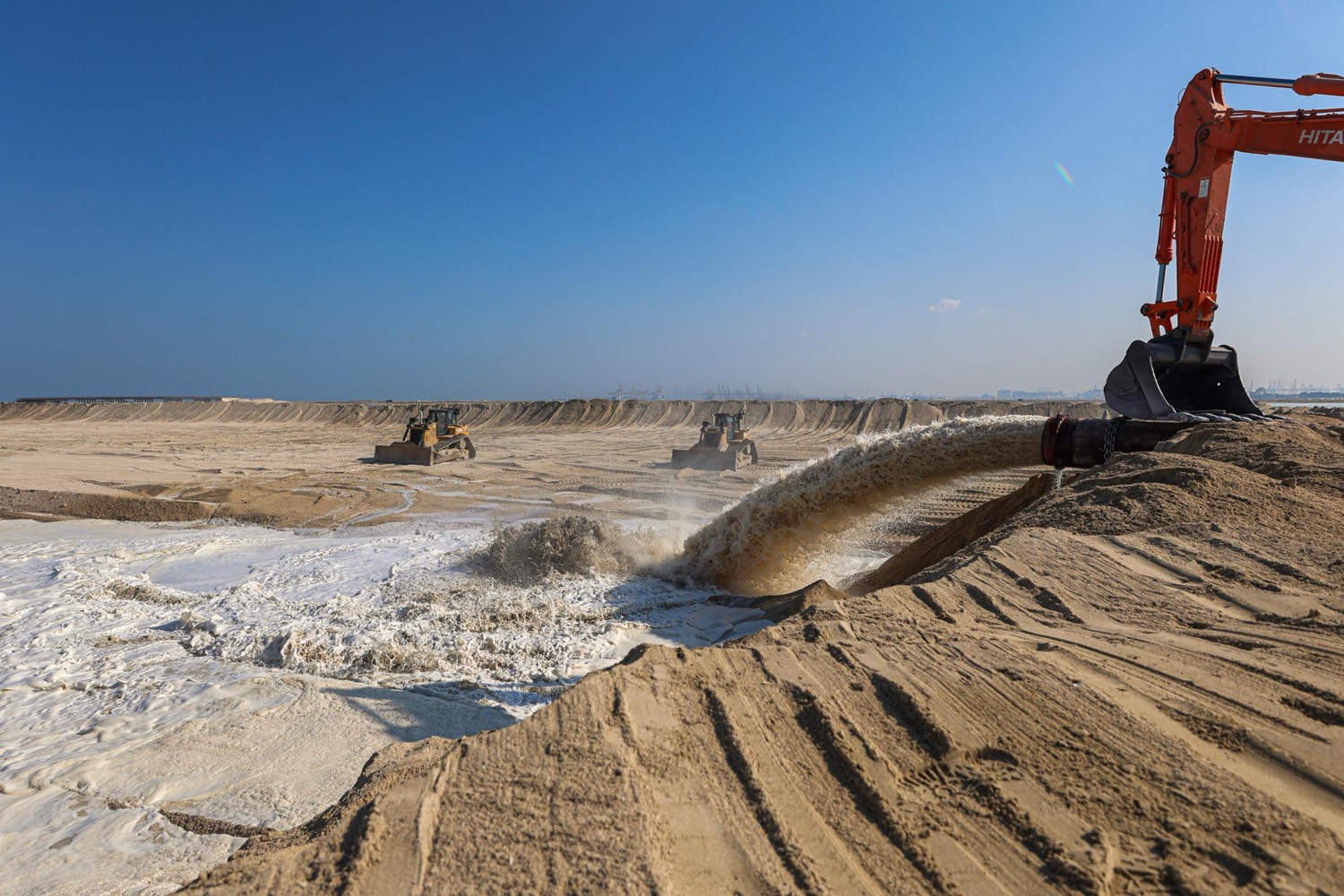 A discharge pipe pumping a torrent of sand and water onto flat reclaimed ground, with bulldozers working in the distance under a clear sky