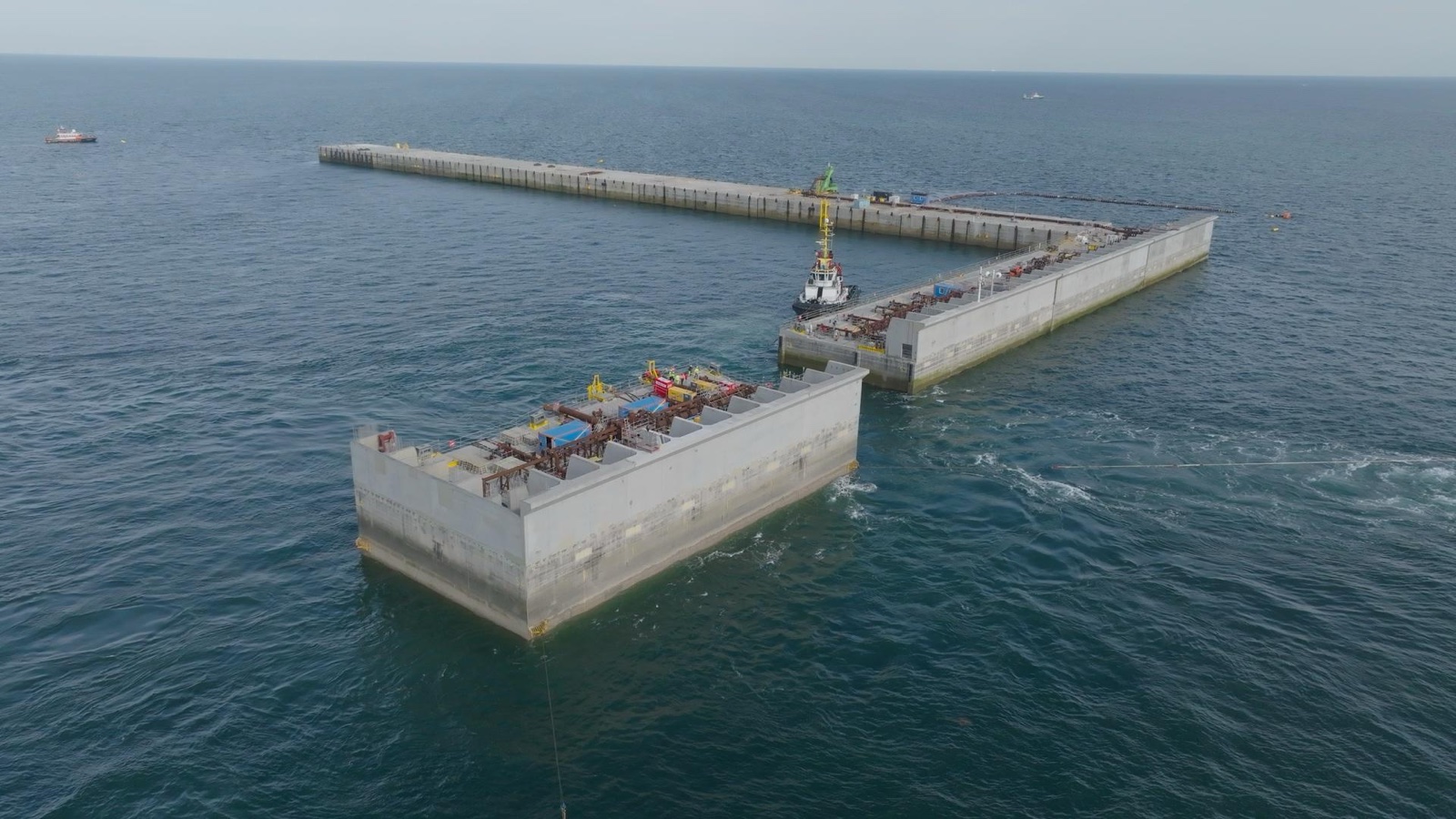 A concrete caisson under tow towards the part-built perimeter wall of Princess Elisabeth Island in the open North Sea, tugs and crew alongside the standing caissons