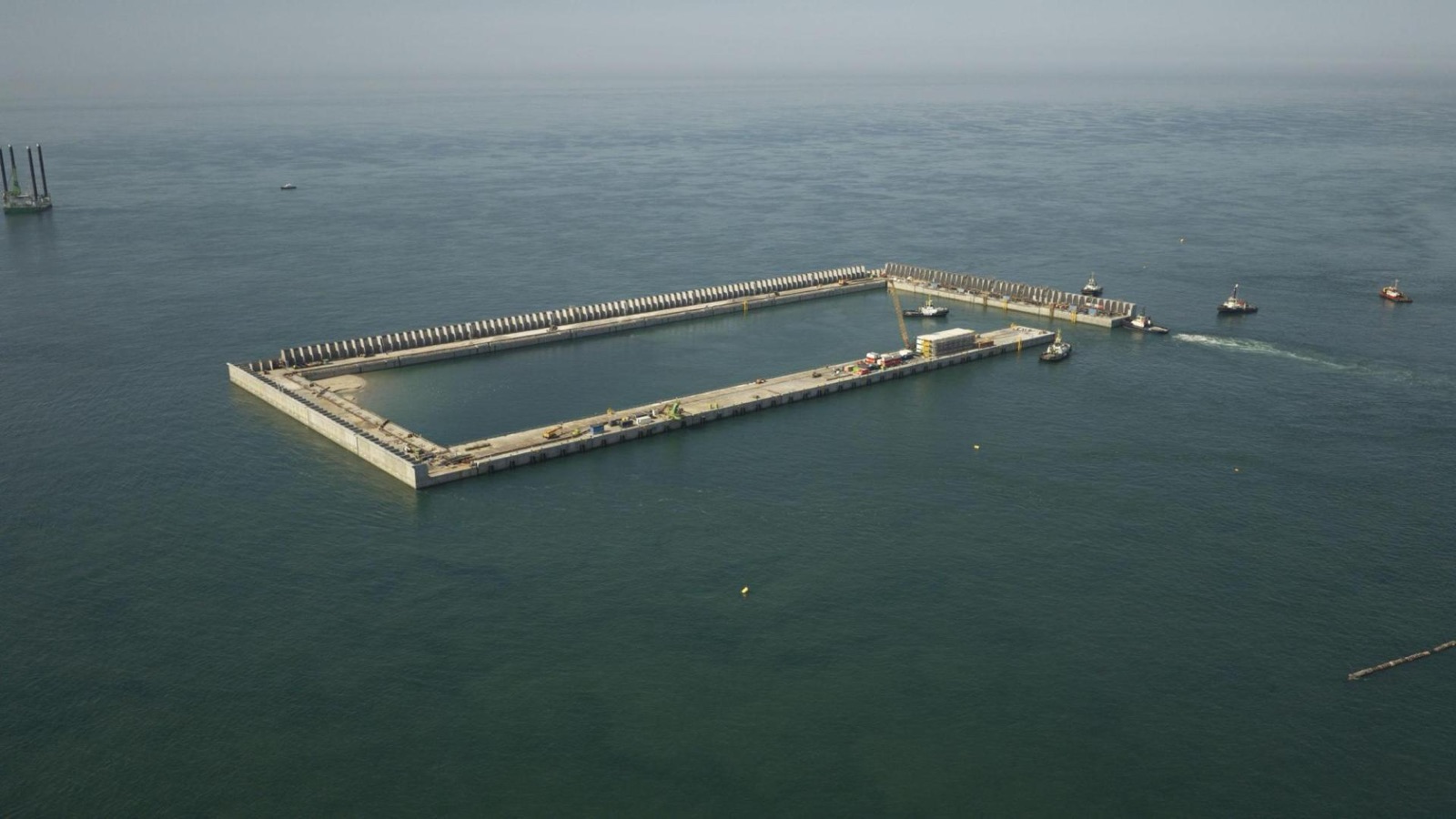 Aerial view of a long rectangle of concrete caissons standing in open sea, its interior still flooded, with tugs towing a caisson towards the last gap in one corner and a jack-up rig on the horizon