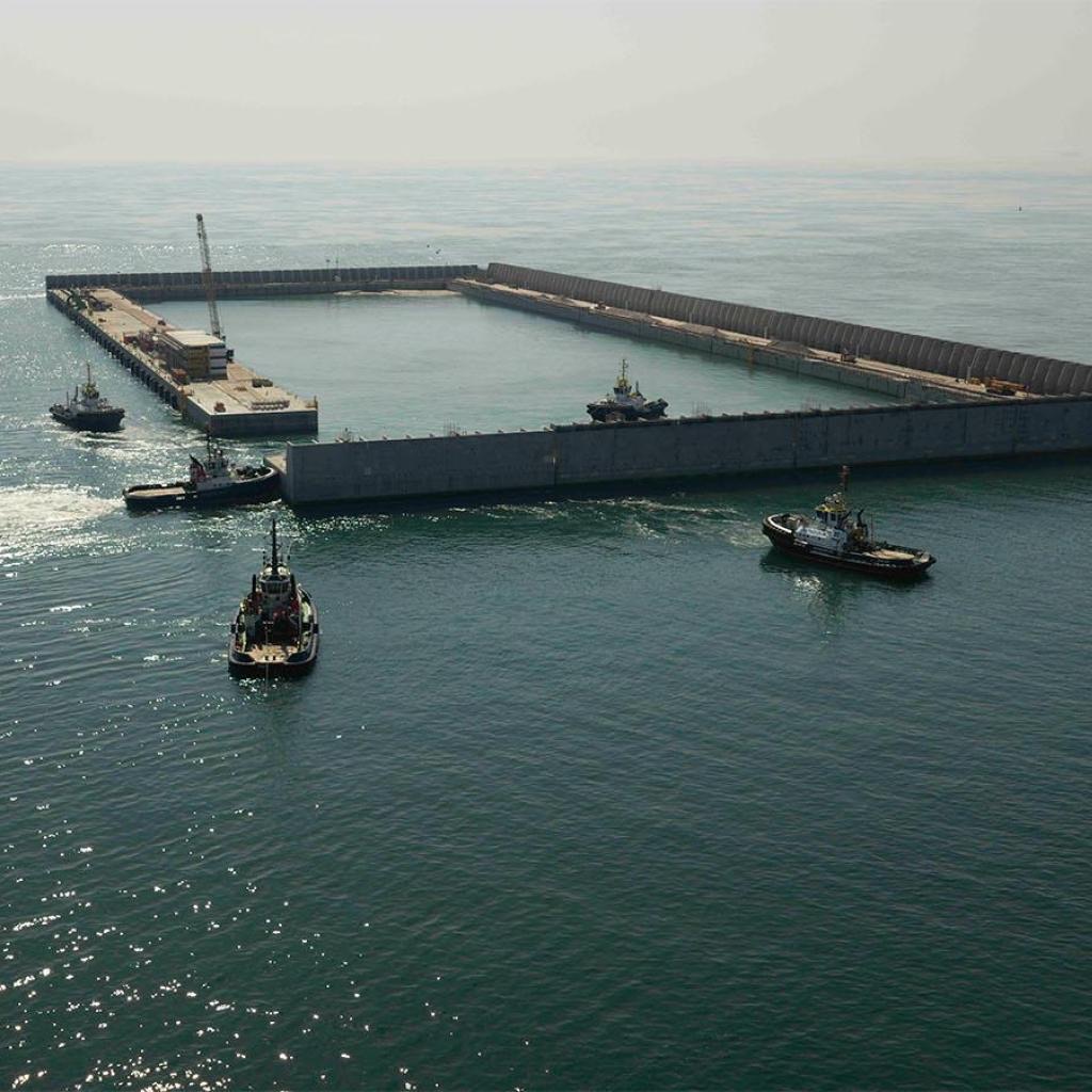 Four tugs manoeuvring a concrete caisson into position at the corner of the island's wall, with a crane and site cabins on the completed caissons behind