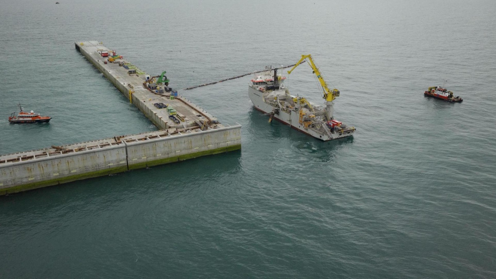 A rock installation vessel with a yellow crane working alongside the concrete wall of the island, a line of floating pipe running between them and two small workboats nearby