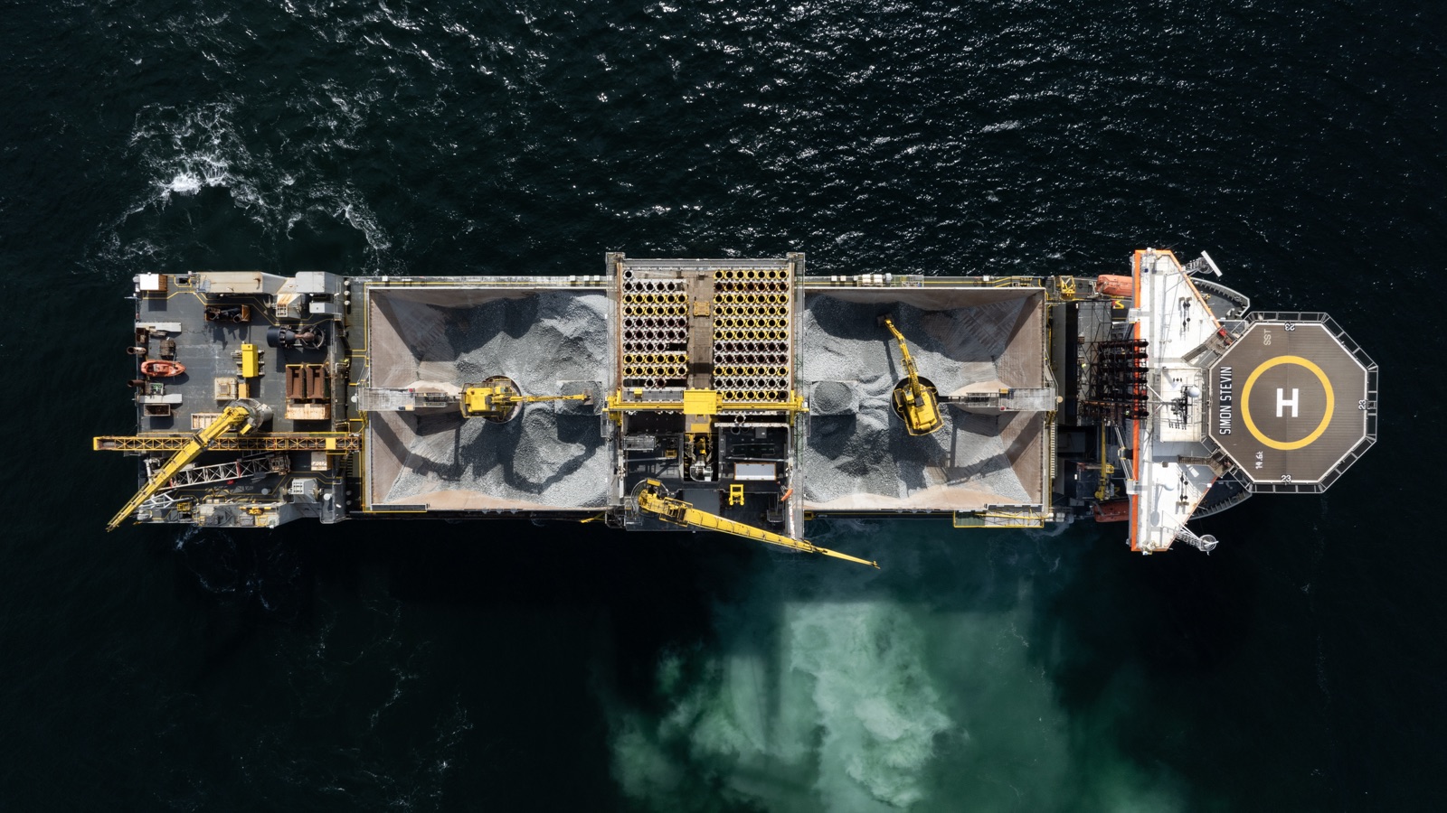 Top-down aerial of the fallpipe vessel Simon Stevin at the island site, rock in its holds and a pale plume in the water where stone is being placed on the seabed