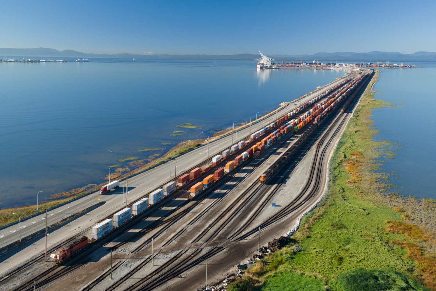 Aerial view of the Roberts Bank causeway: road and rail lines loaded with container trains crossing open water to the man-made terminal island on the horizon