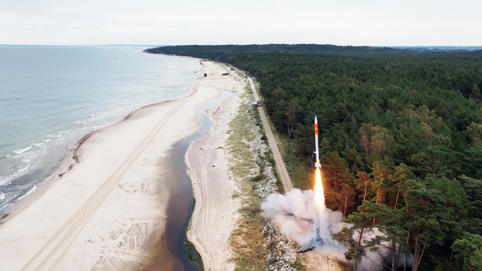 Aerial view of a Perun rocket lifting off from the tree-lined Ustka range on Poland's Baltic coast, its exhaust flame bright against beach and forest