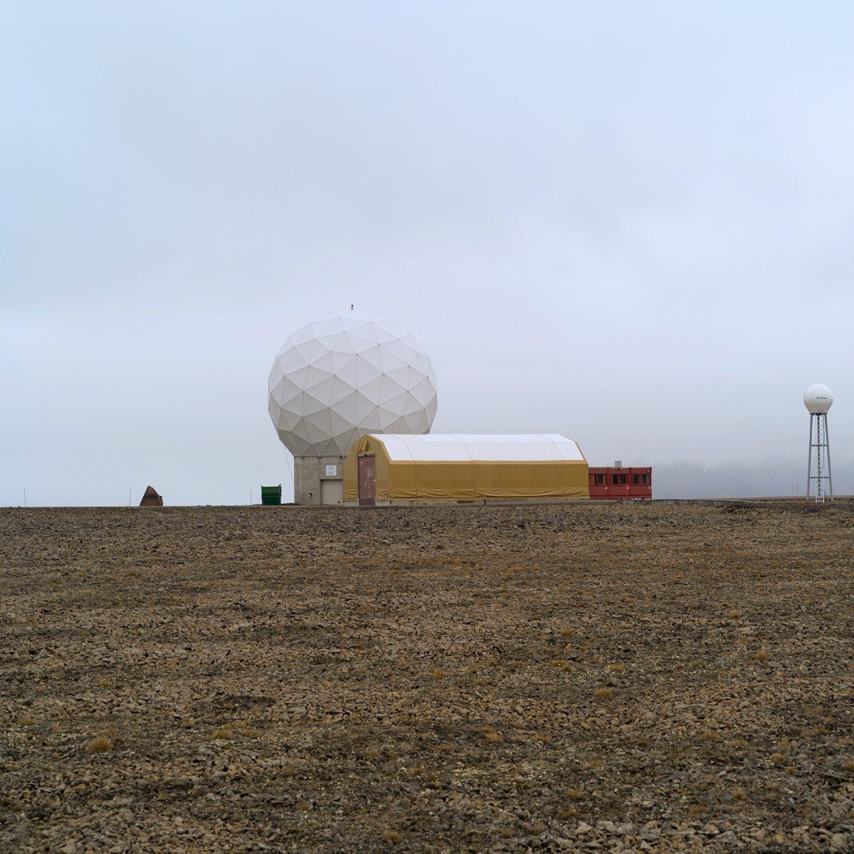A large white geodesic radome and low service buildings standing alone on a bare stony plateau under Arctic cloud